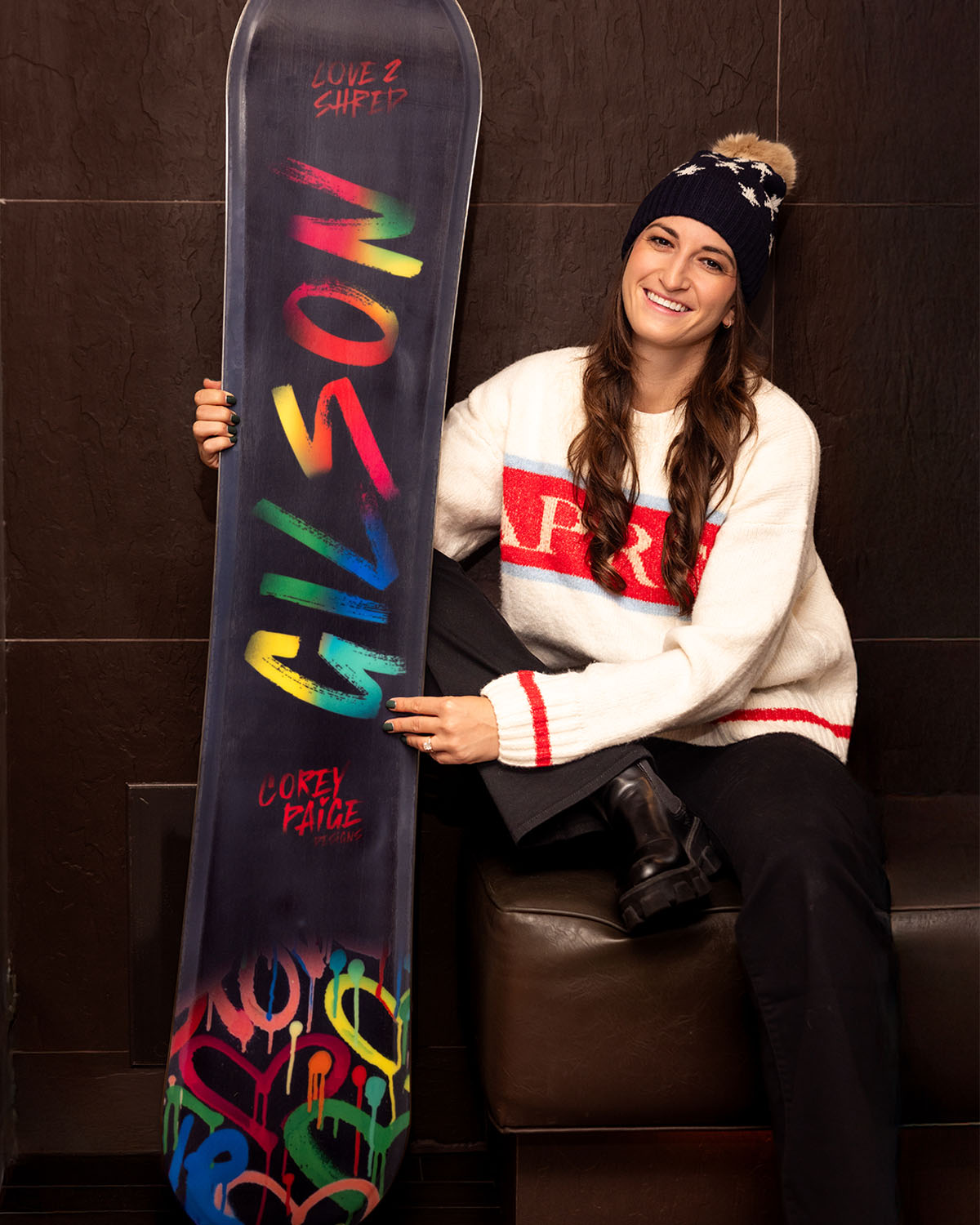 Smiling woman wearing a white sweater with red accents and a navy winter hat sitting indoors, holding a snowboard featuring colorful spray paint-style lettering that reads “GILSON” and “Corey Paige Design.”