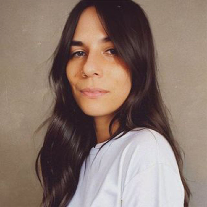 Woman with long dark hair and a nose ring wearing a light-colored shirt, standing against a neutral background with a calm and confident expression.