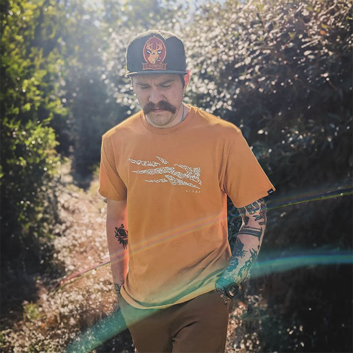 Man walking outdoors on a sunny path wearing an orange graphic t-shirt with a stylized design, a black hat with a deer emblem, and showing tattoos on his arms as light flares cross the image.