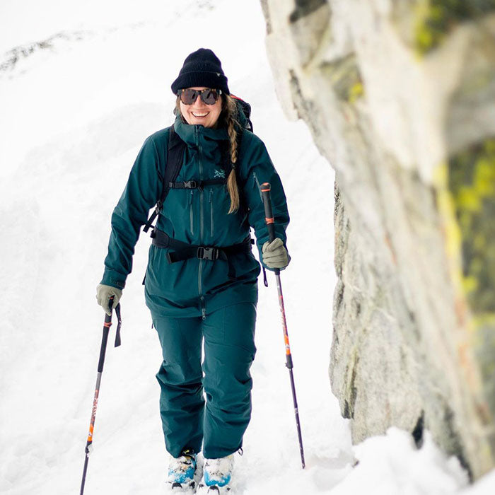 Woman dressed in dark teal winter gear hiking through snowy terrain with ski poles, smiling and wearing sunglasses and a black beanie beside a rocky mountain wall.