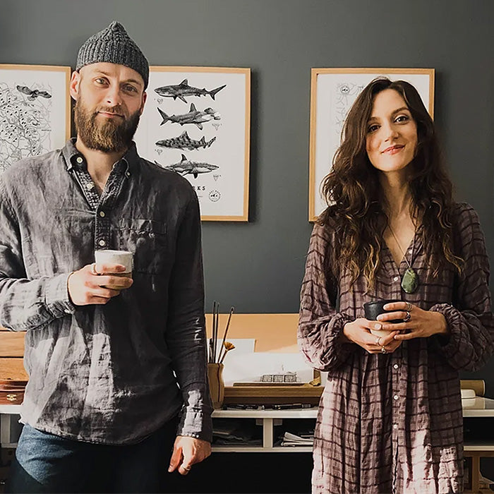 Two artists standing side by side in a warmly lit studio, each holding a cup, with framed illustrations of sharks and maps hanging on the dark gray wall behind them.