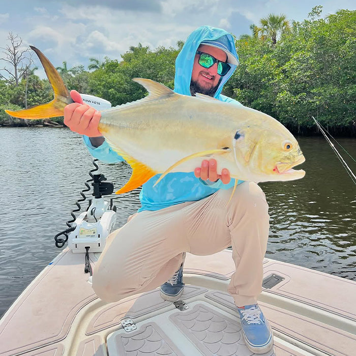 Man wearing a blue fishing hoodie, cap, and reflective sunglasses kneeling on a boat deck while holding a large golden jack fish over calm water with mangroves in the background.