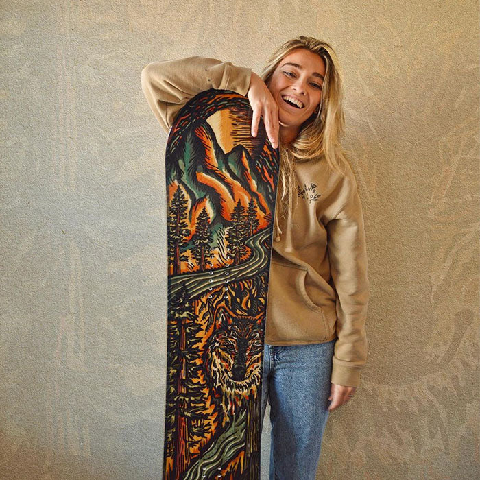 Smiling woman in a tan hoodie and jeans holding a snowboard featuring bold mountain artwork with winding roads, tall pine trees, and a hidden tiger face integrated into the forest design.