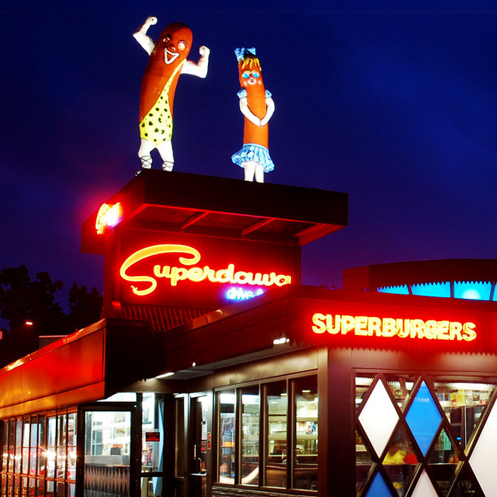 Retro style diner at night with bright red neon signs reading “Superdawg” and “Superburgers,” topped by two giant hot dog mascots, one flexing and the other wearing a skirt, glowing against a deep blue sky.