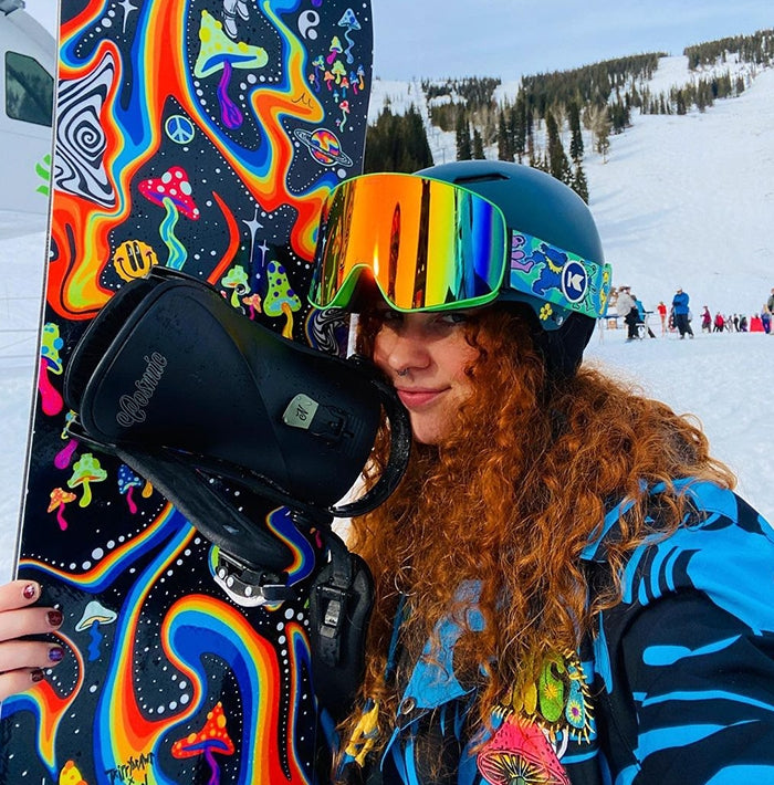 Snowboarder holding a board covered in colorful psychedelic artwork with swirling rainbow patterns, mushrooms, and space motifs, wearing a rainbow-lens helmet and blue jacket on a snowy mountain slope.