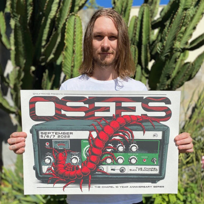 Man standing outdoors in front of tall green cacti holding a concert poster for the band OSEES, featuring bold red lettering and a large red centipede crawling across a green amplifier design.