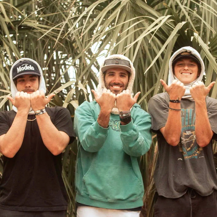 Three friends standing in front of tall palm leaves wearing casual hats and shirts, each smiling and holding up shaka hand signs in a relaxed, tropical setting.