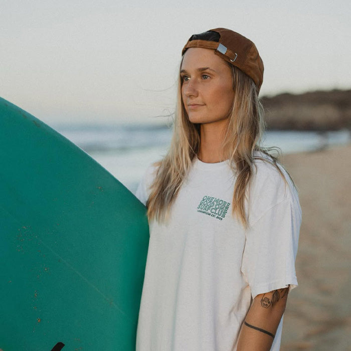 Woman standing on a sandy beach holding a teal surfboard, wearing a white t-shirt with a small graphic and a brown backward cap, looking toward the ocean in soft evening light.