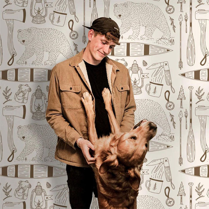Man wearing a tan jacket and backward cap smiling while holding a golden retriever, standing in front of a patterned wall featuring bears, canoes, axes, mugs, and other outdoors-themed illustrations.