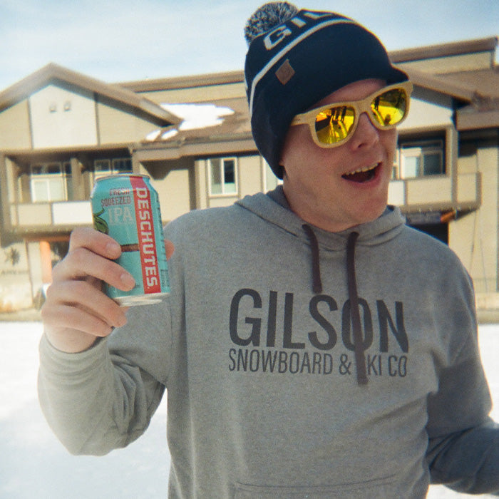 Man wearing a Gilson Snowboard and Ski Co hoodie, matching winter hat, and reflective sunglasses, smiling and holding a can of Deschutes Fresh Squeezed IPA outdoors in a snowy mountain town setting.